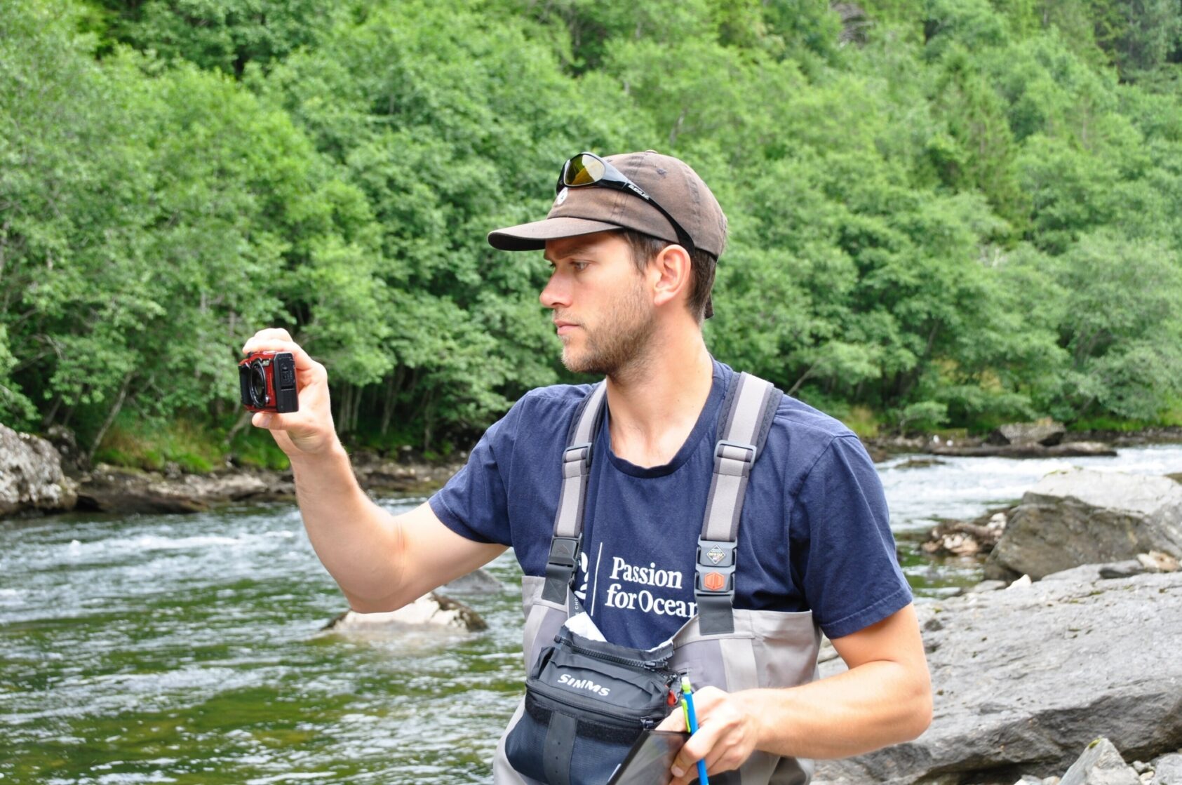 Hallvard Tysse, Samningen, NORCE-forsker Erlend Mjelde Hanssen jakter svar ved hjelp av sensorer og lytteutstyr. Drepes laksesmolten i Modalen og Tysse av turen gjennom rørgater og kraftturbiner? Foto: Hallvard Tysse, Samningen, Erlend foto Hallvard Tysse Samningen, <p>Hallvard Tysse, Samningen</p>, Mann i vadere fotograferer med et lite kamera
