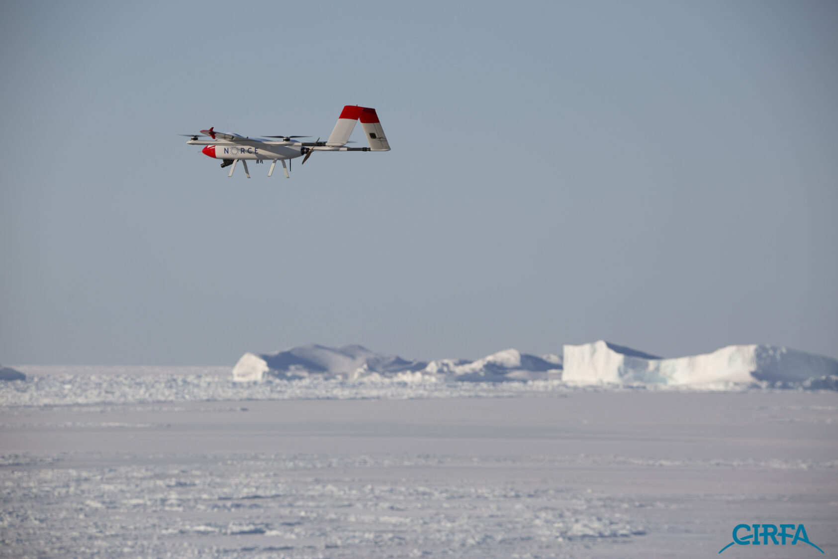 Foto: William Copeland/MET Norway, Baby Shark er en fastvingedrone med et vingespenn på 2,5 meter. Dronen er utstyrt med kameraer som gir automatisk eksponeringskontroll og legger til georeferanse på bildene i sanntid før den deler bildene med bakkestasjonen og nLive-serveren., William Copeland 2, <p>William Copeland</p>, En drone flyr over isen. Isfjell i bakgrunnen.