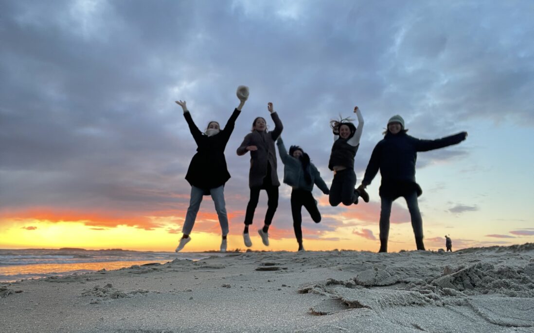 Fem unge glade mennesker med hopper luften på en stranden. Solnedgang i bakgrunnen.