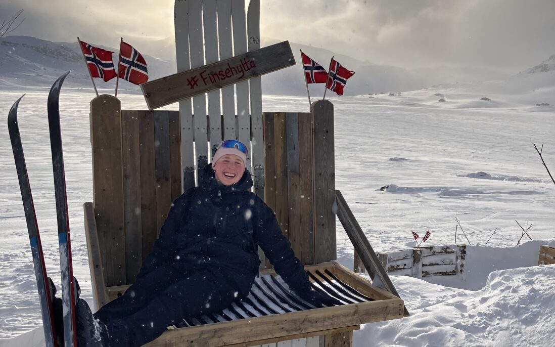Kvinne med ski på beina sittende på en trebenk ute i snøen.