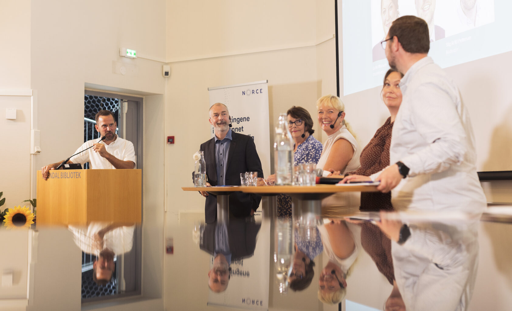 Andreas R. Graven, Våre arrangementer foregår på biblioteket i Arendal, her fra debatten i 2023., Lege-vakt-debatt-jesper-arendalsuka-23, <p>Andreas Graven, NORCE</p>, Fem personer bak runde bord, og en debattleder bak en talerstol. Personene speiles gså i en flate i forgrunnen.