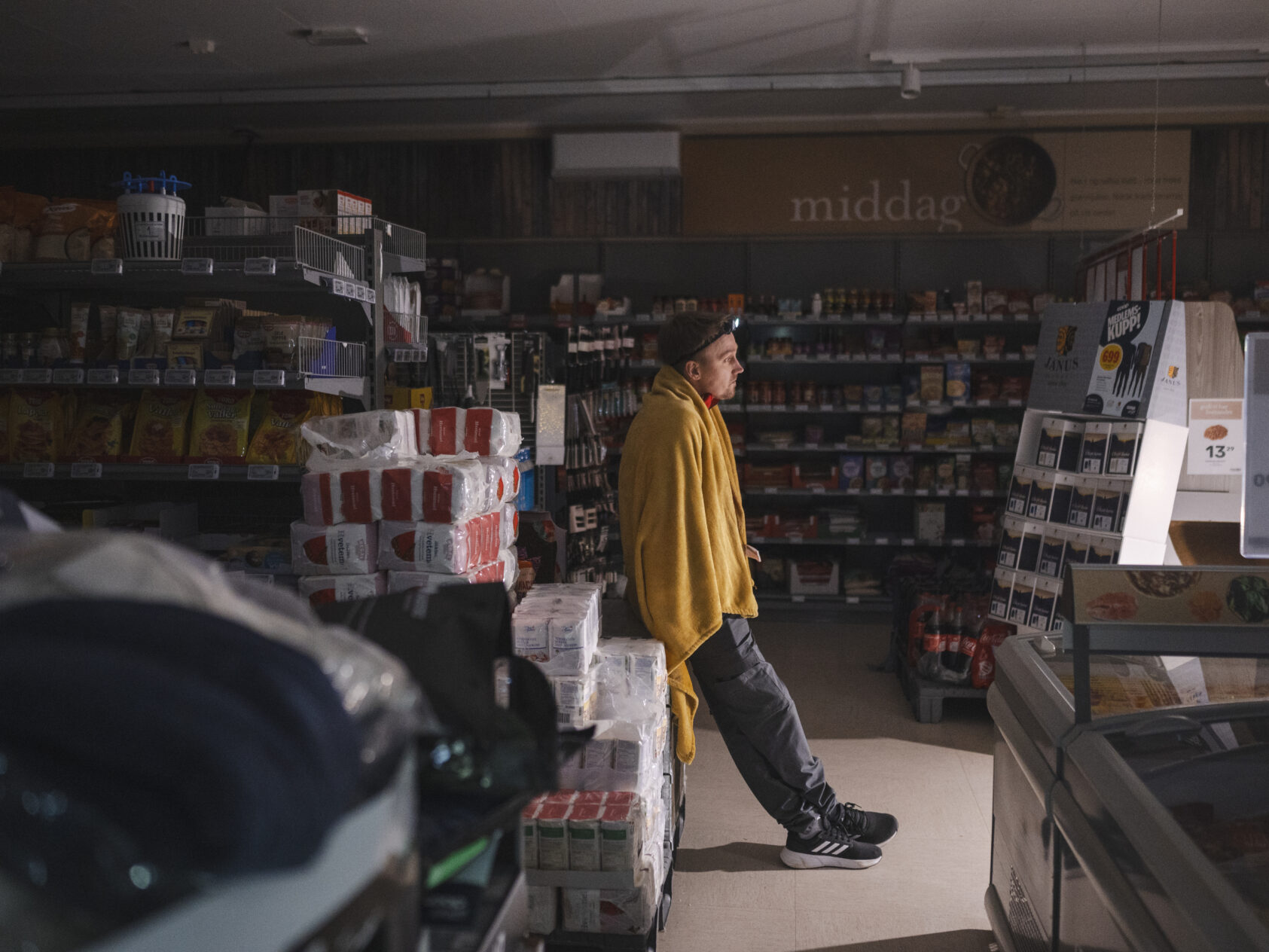 , Simen Kristiansen forteller om en kald og stille arbeidsdag i en strømløs butikk i Kvam i Trøndelag i januar 2025. Foto: Sebastian Bang, Steinkjer Avisa, Illustrasjon, <p>Sebastian Bang</p>, Mann lener seg mot en butikkhylle, mørkt i rommet