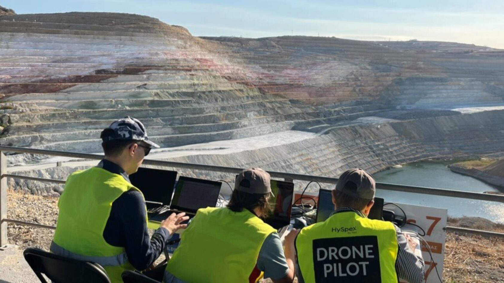 NEO, Dronepiloter flyr drone med hyperspektral kamera fra Norsk Elektro Optikk AS. Vi samlet data fra en bentonittgruve i Hellas for EU-prosjektet M4Mining. På bildet ser du (f.v.) Axel Fredrik Bohman, Friederike Körting og Justus Constantin Hildebrand, alle fra NEO., M4minig NEO, <p>NEO</p>, Tre personer sittennde ute foran bord med pc og annet utstyr, åpen gruve i bakgrunnen
